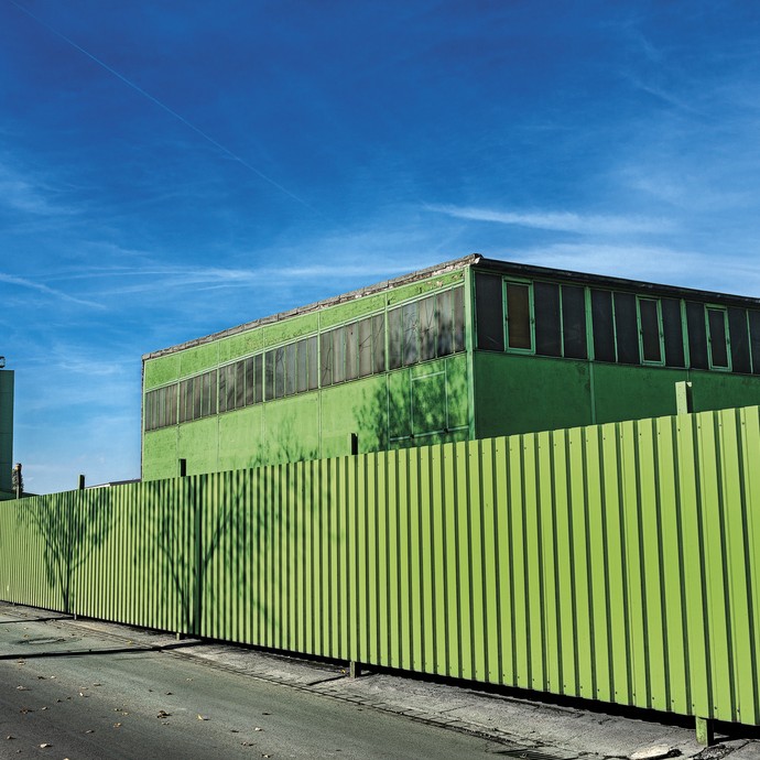 Ein grasgrünes Fabrikgebäude vor einem tiefblauen Himmel, Schatten verschiedener Bäume sind auf der Umzäunung der Fabrik zu sehen (vergrößerte Bildansicht wird geöffnet)