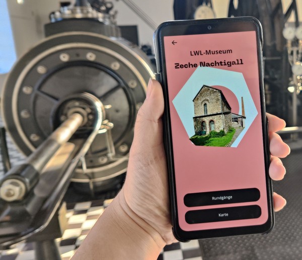 Smartphone mit dem Startbildschirm der App. Im Hintergrund die Dampffördermaschine.
