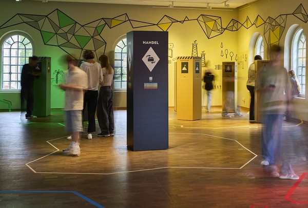 Blick in den Ausstellungsraum. Im Mittelpunkt eine blaue Stele mit dem Begriff Handel. Schülerinnen und Schüler sind in Bewegungsunschärfe darstellt.