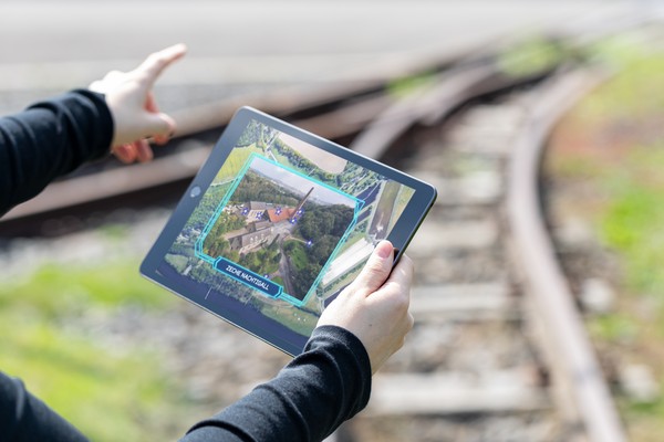 Hände halten ein Tablet und im Hintergrund sind die Gleise der Zeche Nachtigall zu sehen.