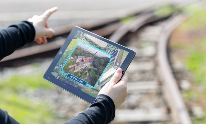 Blick auf ein Tablet in den Händen einer Besucherin. Im Hintergrund sind Gleise der Zeche Nachtigall zu sehen.
